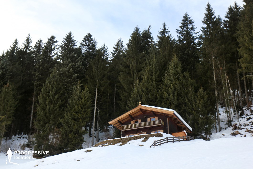 This serene mountain location features a traditional alpine wooden cabin nestled at the edge of a dense pine forest. The dark wooden structure, with its gabled roof and balcony, stands in contrast to the snow-covered ground and tall evergreen trees that dominate the background. The snow adds texture and brightness to the foreground, while the vertical rhythm of the trees creates a natural frame around the building. This setting offers a secluded, atmospheric backdrop ideal for winter scenes, remote retreat sequences, or productions requiring a picturesque forest lodge ambiance.
