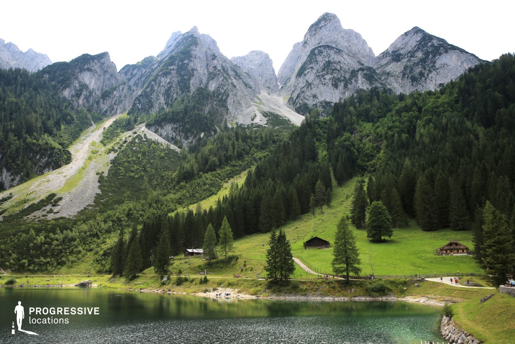 This striking landscape features a glassy mountain lake bordered by a gently sloping green meadow dotted with rustic wooden lodges, all set against a dramatic backdrop of steep, rocky peaks. The vivid contrast between the lush lower terrain and the stark alpine cliffs creates a visually rich and immersive setting. Ideal for fantasy epics, romantic dramas, or adventure films, this location is perfect for scenes of arrival, peaceful retreat, or the beginning of a mountainous journey.