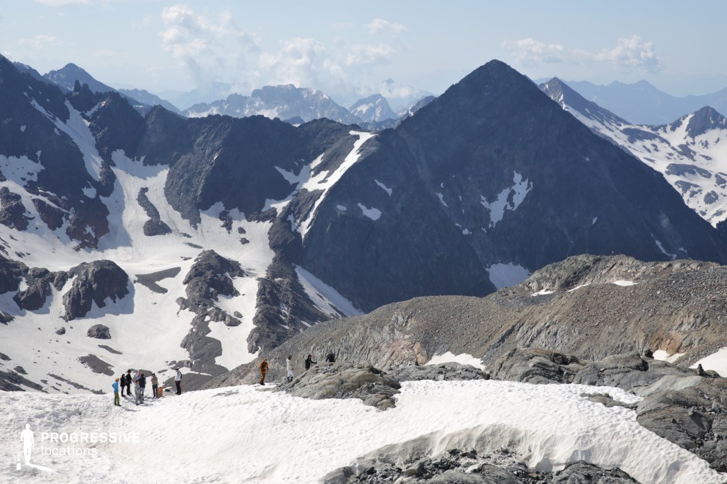 This high-altitude location showcases a dramatic, snow-covered alpine expanse framed by rugged dark peaks and patches of glacial ice. A group of hikers in the foreground adds scale and human presence to the otherwise vast and untouched environment. The contrasting textures of snow, rock, and distant ridgelines make this scene ideal for epic sequences, expedition-style storytelling, or dramatic establishing shots set in extreme natural environments. Suitable for fantasy, survival, or adventure genres where nature plays a dominant visual and thematic role.