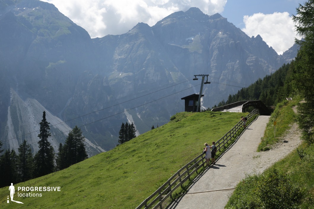 Set on a gently sloping grassy ridge, this location features a panoramic view of towering rocky peaks layered in misty gradients. A narrow mountain path runs alongside a rustic wooden fence, with a small cable car station and support pole adding subtle structural interest. The combination of natural grandeur and human-scale detail makes this site well-suited for romance, drama, or travel films—particularly for scenic contemplative sequences, emotional turning points, or characters pausing during a journey through the mountains.