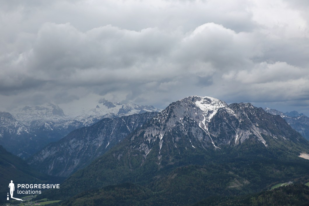 This sweeping mountain vista showcases a rugged alpine range with sharp, snow-dusted peaks rising from dark forested slopes, layered against a backdrop of even higher, glacier-covered summits. The heavy cloud cover and deep valleys create a sense of grandeur and isolation, enhancing the raw, untamed atmosphere. This location is ideal for epic fantasy films, survival thrillers, or historical adventures, especially scenes involving treacherous journeys, ancient strongholds, or moments of awe in the face of nature’s overwhelming scale.