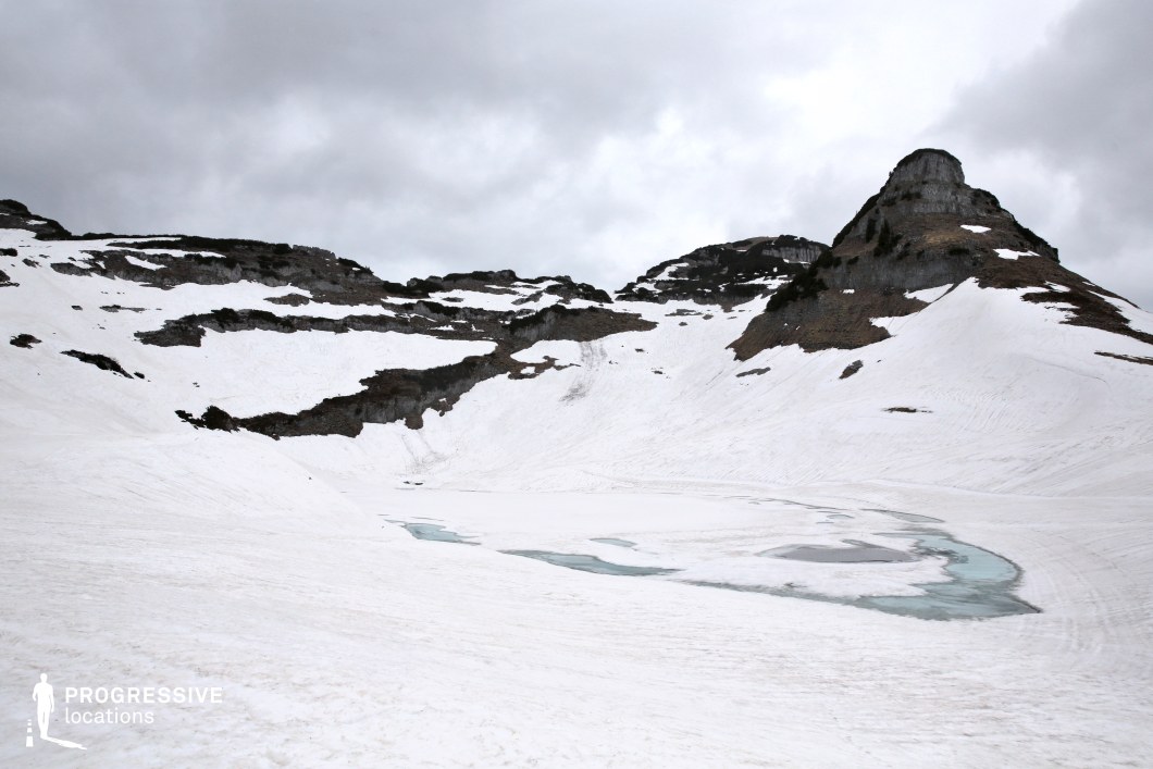 This stark, high-altitude landscape presents a wide snow-covered basin with patches of ice and meltwater forming abstract shapes around its center. Jagged rock formations emerge from the snow on all sides, while a single prominent peak adds vertical focus. The minimal color palette and harsh terrain lend the location a raw, otherworldly aesthetic. Perfect for filming scenes set in remote or extreme environments, this location suits survival stories, fantasy journeys, or science fiction worlds that require a sense of isolation, peril, or untouched wilderness.