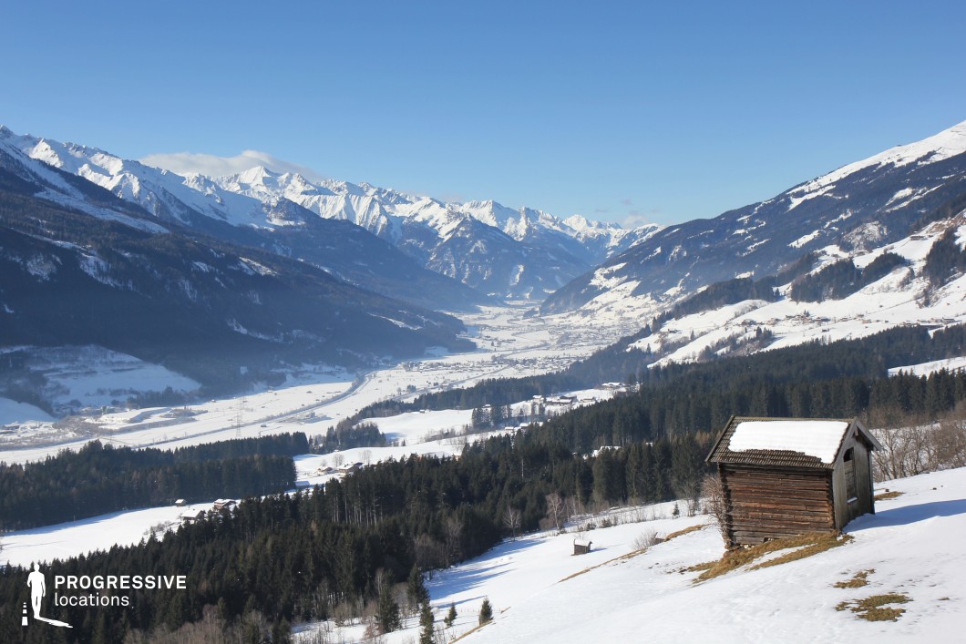 This expansive alpine landscape features a snow-blanketed valley flanked by towering, rugged mountain ranges. A solitary wooden hut perched on a gentle slope adds rustic character and a human scale to the otherwise vast and untamed panorama. The layering of forest, open fields, and jagged peaks creates a compelling visual depth. This location is ideal for period dramas, epic journeys, or winter-themed productions—particularly scenes involving isolation, introspection, or cross-country travel through a majestic and remote environment.