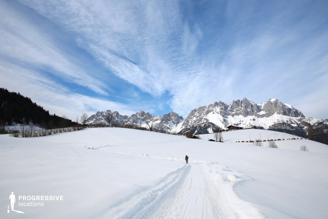 This wide-angle image captures a serene snow-covered expanse framed by imposing, jagged alpine peaks in the distance. The gently sloping white terrain is uninterrupted, emphasizing the grandeur and scale of the natural surroundings. A snow-covered path leads the viewer’s eye toward the dramatic rock formations, which rise boldly from the undulating hills. The dark tree line on one side provides contrast to the vast, luminous snowfield, enhancing the visual depth and balance of the composition. This location is ideal for filming due to its unspoiled, cinematic quality and strong natural contrast. It lends itself perfectly to genres such as drama, adventure, fantasy, and historical epics—ideal for scenes of isolation, journeys, or transformative moments in nature.