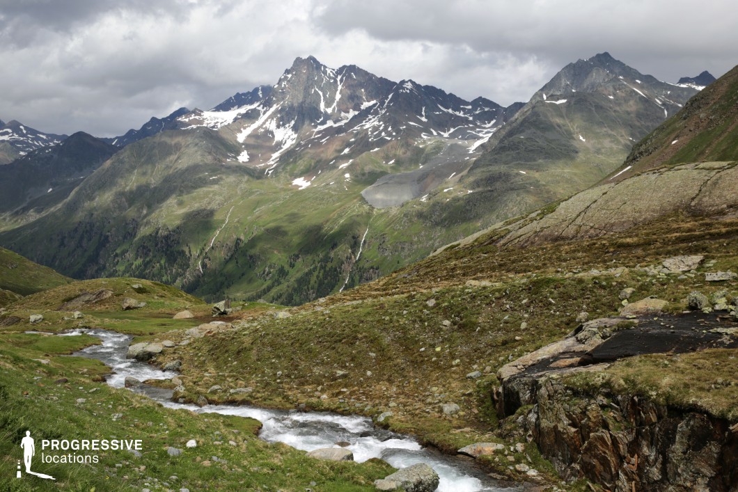 This majestic mountain setting features a lively alpine stream winding through a green, rocky highland plateau, with towering snow-streaked peaks rising dramatically in the background. The landscape transitions from soft meadows to rugged heights, offering both intimacy and grandeur. The location is perfect for adventure epics, historical dramas, or fantasy tales—ideal for scenes involving travel, escape, or moments of clarity in nature’s vastness.
