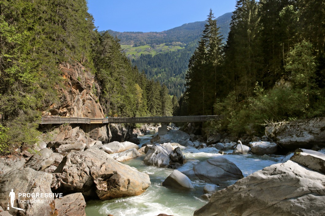 This dramatic valley scene showcases a turbulent mountain stream coursing through a narrow gorge filled with large boulders, framed by steep rock walls and towering coniferous forest. A wooden bridge spans the gap above the water, providing both a visual anchor and a cinematic passage point. The natural contrast between rock, water, and forest creates a dynamic environment perfect for action sequences, escape scenes, or moments of revelation in adventure films, fantasy epics, or nature-centric dramas.