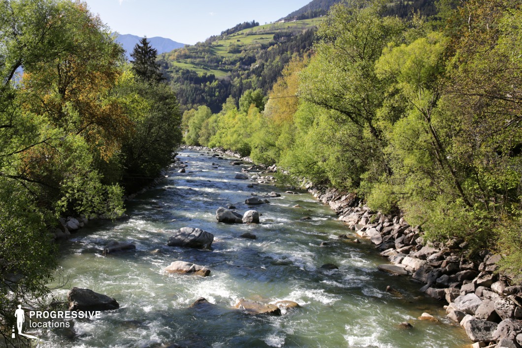 his dynamic landscape features a swift-flowing river weaving through a narrow, tree-lined valley, with boulders breaking the current and forested slopes rising toward distant hills. The dense greenery, rocky banks, and layered hillsides create a vibrant natural corridor rich in texture and depth. Ideal for adventure films, period dramas, or fantasy stories, this location is perfect for river journeys, wilderness crossings, or scenes of escape, confrontation, or discovery in a lively and immersive mountain setting.