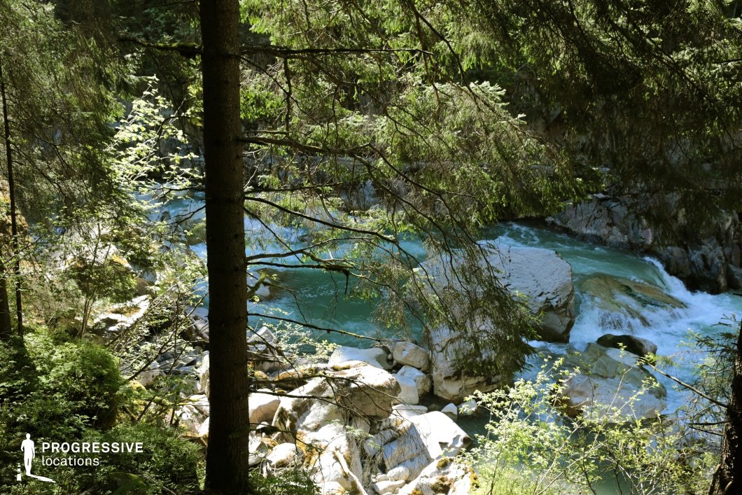 This location features a vibrant turquoise stream cutting through a forested gorge, with water flowing around large white boulders and framed by tall evergreen trees. The interplay of light and shadow, along with the contrast between the rushing water and still pools, creates a dynamic and immersive natural setting. Ideal for fantasy, survival, or adventure films, this scene works perfectly for moments of discovery, refuge, hidden crossings, or dramatic natural challenges encountered deep within the wilderness.