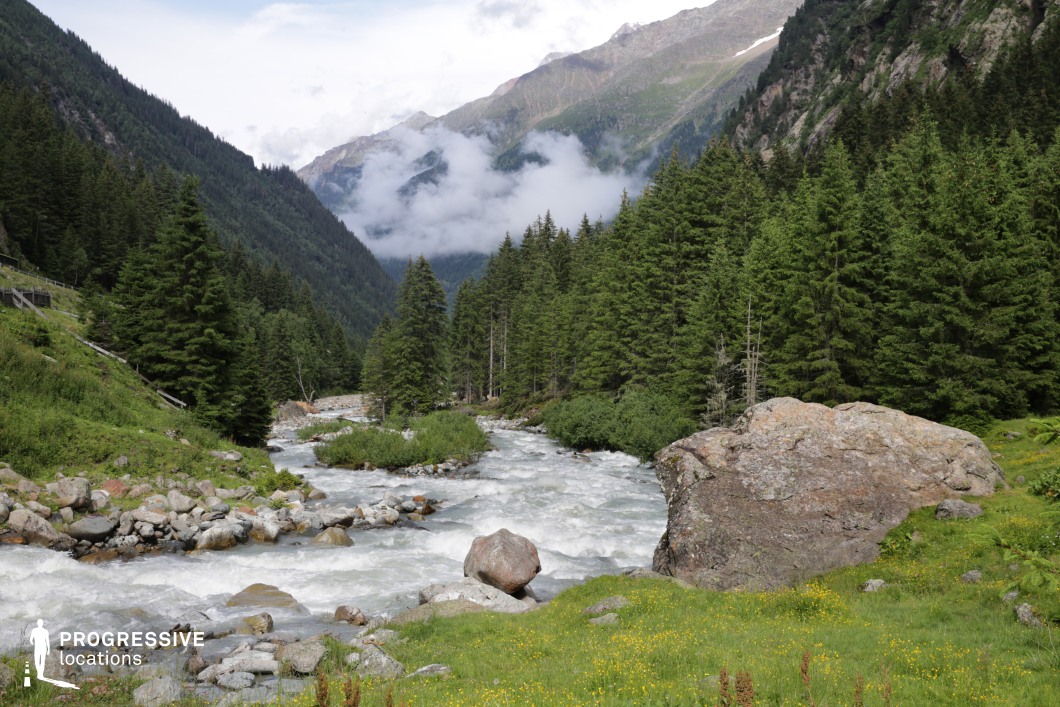 A powerful alpine river cuts through this deep valley, flanked by rugged boulders, wild meadows, and dense pine forest, all enclosed by towering mountain slopes. The layering of natural elements and the dramatic convergence of terrain create a cinematic sense of movement and scale. This location is ideal for epic outdoor adventures, survival stories, or fantasy quests—particularly scenes of wilderness travel, perilous crossings, or moments of discovery within a majestic and untamed environment.