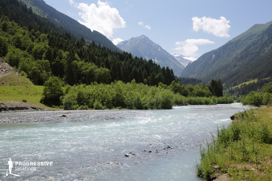 This stunning mountain valley features a clear, fast-flowing river winding between densely forested slopes and sharp alpine peaks. The lush greenery and snow-capped mountains in the distance provide a dramatic yet serene natural contrast, creating a visually rich and immersive setting. This location is ideal for epic adventure films, fantasy journeys, survival stories, or nature documentaries, offering a versatile backdrop for both sweeping landscapes and intimate riverside scenes.