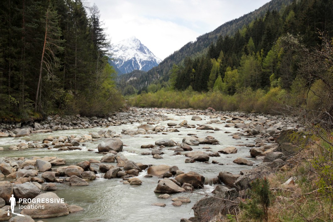 This striking mountain valley features a wide, boulder-strewn river coursing through densely wooded slopes, framed by coniferous forests and culminating in a snow-capped peak in the distance. The milky, glacial water winds through the uneven terrain, with smooth rocks breaking the flow and adding texture to the riverbed. The scene is layered with rich natural elements—stone, water, trees, and elevation—creating a dynamic yet balanced visual environment. Ideal for outdoor adventure, survival, or nature-focused films, this location offers a dramatic blend of movement and stability, making it suitable for both action and introspective sequences in a pristine alpine setting.