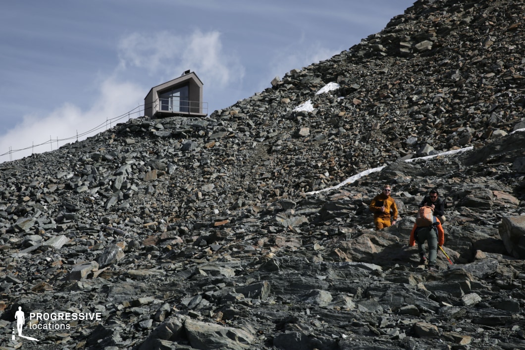 A steep, desolate slope of jagged rocks leads the eye to a small, angular shelter perched near the crest, surrounded by a minimal safety railing. The sparse patches of snow and sharp rock textures create an inhospitable yet striking environment, enhanced by the structure’s modern, geometric form. The remote setting and dramatic elevation are ideal for survival thrillers, sci-fi dramas, or introspective stories involving isolation, endurance, or the encounter with the sublime in a harsh natural landscape.