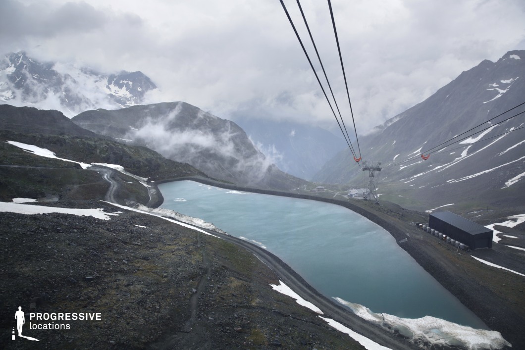 This elevated artificial lake is nestled within a stark, glacial mountain landscape, partially edged with snow and mist, and surrounded by steep slopes and rugged paths. The scene is dominated by a cableway system, industrial structures, and a desaturated color palette, creating a remote, otherworldly atmosphere. The stark isolation and raw textures make this location ideal for science fiction, survival, or thriller genres—particularly scenes involving secret facilities, extreme expeditions, or environmental crises at the edge of civilization.