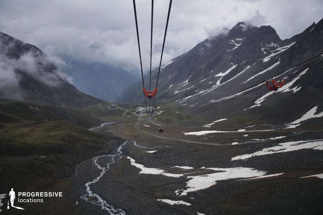 This expansive high-altitude scene is defined by steep, rocky slopes, patches of lingering snow, and a winding stream cutting through the valley floor, all intersected by a suspended cable car system. The towering mountains and misty atmosphere enhance the grandeur and isolation of the terrain, while the infrastructure adds a modern, adventurous element. This location is ideal for action films, mountaineering dramas, or thrillers involving pursuit, survival, or escape, especially for scenes set in remote outposts, hidden facilities, or perilous highland crossings.