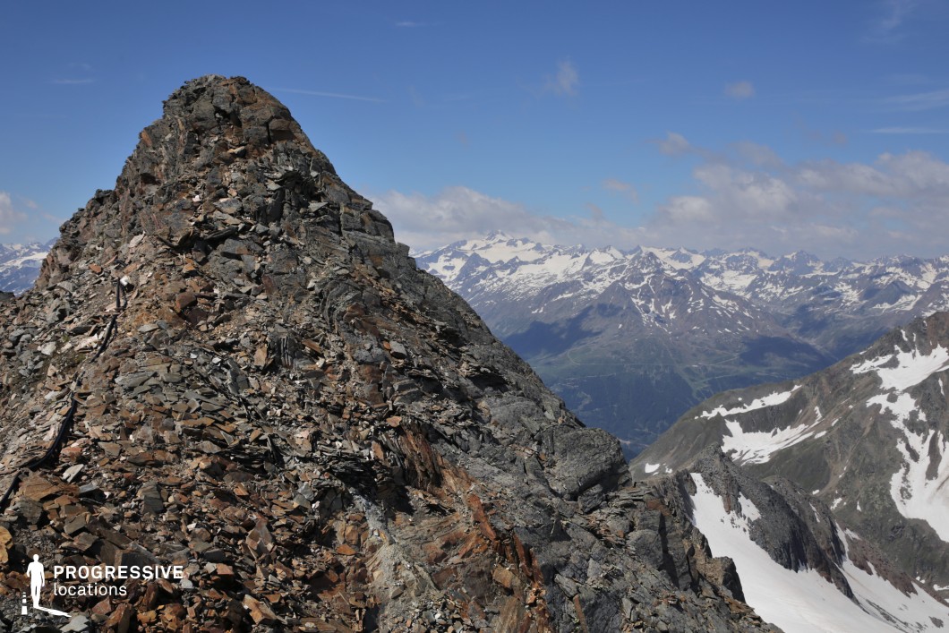 This dramatic rocky peak rises sharply, composed of broken shale and rugged formations, with sweeping views of snow-dusted mountain ranges stretching into the distance. The stark contrast between the jagged foreground and the vast, layered alpine landscape enhances the sense of scale and remoteness. This location is ideal for filming epic adventure, survival, or fantasy scenes involving mountain ascents, pivotal confrontations, or moments of awe in extreme, high-altitude environments.