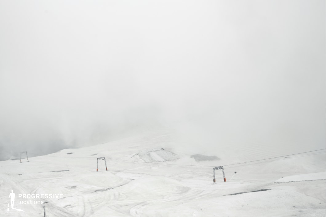 This desolate snow-covered plateau fades into thick fog, with only the silhouettes of ski-lift structures and faint terrain lines breaking the monochrome expanse. The minimalism and soft gradients create a haunting, otherworldly atmosphere, blending natural and industrial elements into a visually abstract space. This location is ideal for psychological thrillers, dream sequences, survival stories, or science fiction films that require isolation, disorientation, or a sense of crossing into the unknown.