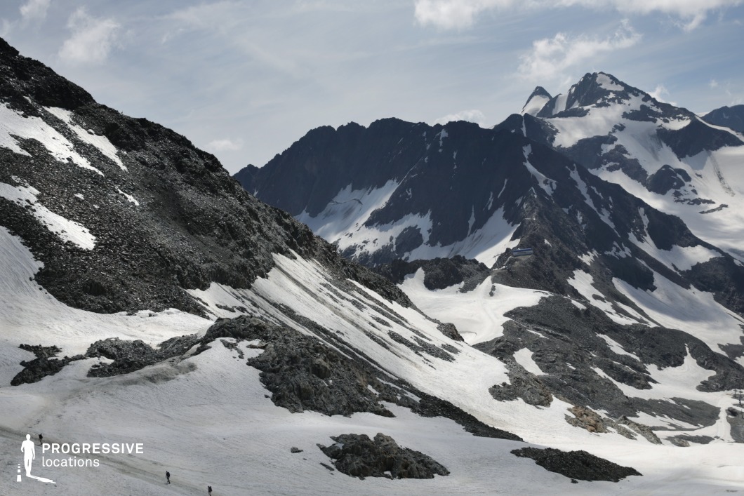 This dramatic high-altitude landscape features a rugged expanse of snow-covered slopes and jagged rock formations, framed by towering black-and-white peaks in the background. The terrain alternates between smooth glacial surfaces and angular, fragmented rock, creating strong contrasts in texture and tone. The deep valleys and high ridges form a dynamic topography, emphasizing isolation and verticality. With its raw, untouched atmosphere and expansive visual depth, this location is highly suited for adventure, survival, or epic genre productions, offering a powerful backdrop for scenes of endurance, exploration, or dramatic confrontation in extreme natural conditions.