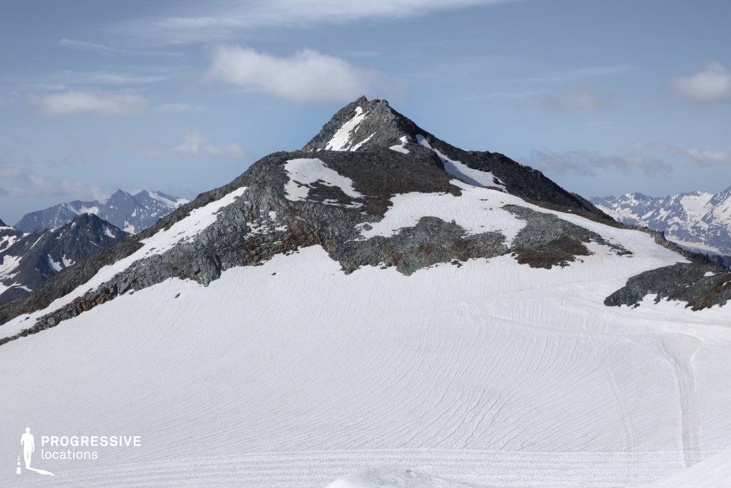 This pristine alpine setting showcases a rugged peak emerging from a broad expanse of compact snow, with dark rock formations breaking through the white surface. The dramatic elevation and untouched terrain offer a stark and powerful visual, surrounded by distant mountain ranges under a clear sky. This location is ideal for high-stakes scenes in survival, action, or fantasy genres—especially summit sequences, harsh wilderness treks, or symbolic moments of conquest, reflection, or isolation.