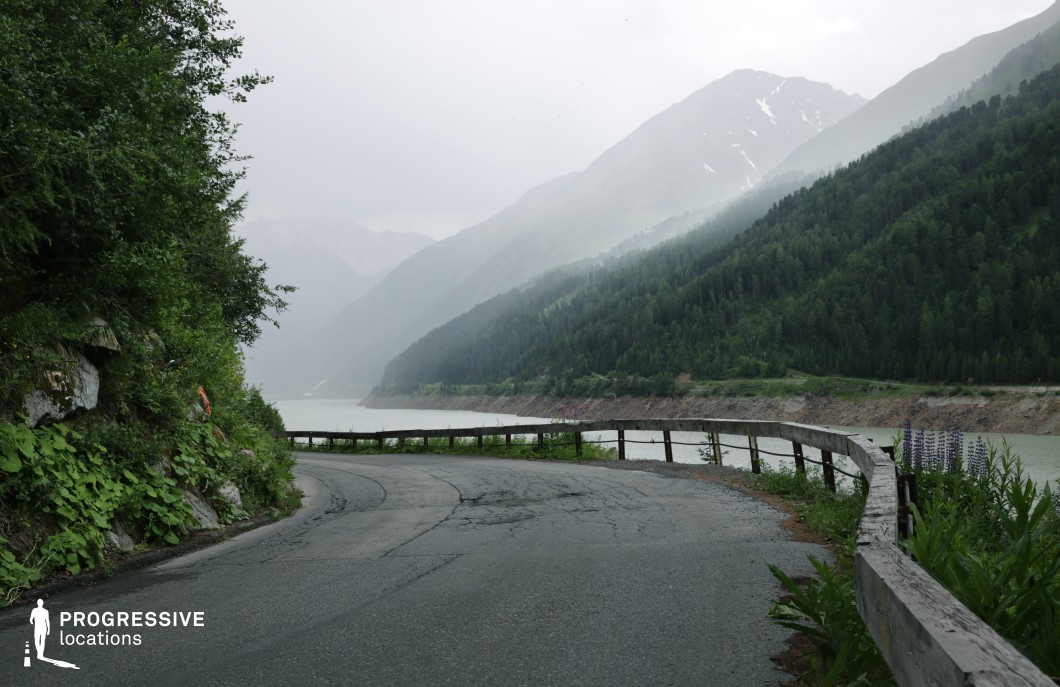 This image captures a winding mountain road bordered by a weathered wooden guardrail, overlooking a large, serene lake flanked by steep forested slopes and distant, rugged peaks. The asphalt surface shows signs of wear, adding to the road’s rustic, natural character. Dense greenery grows along the rocky roadside on the left, while on the right, the railing separates the road from the drop toward the lake, emphasizing the elevation and curvature of the route. The landscape features a harmonious blend of earthy tones, deep greens from the coniferous trees, and the muted blues and greys of the lake and mountains, creating a tranquil yet dramatic visual composition. The distant hills fade into atmospheric perspective, enhancing the sense of depth and isolation. This location is ideal for cinematic scenes requiring a remote or introspective natural backdrop—especially effective for drama, thriller, or travel-themed productions—and offers strong visual contrast for aerial or tracking shots along the winding road.
