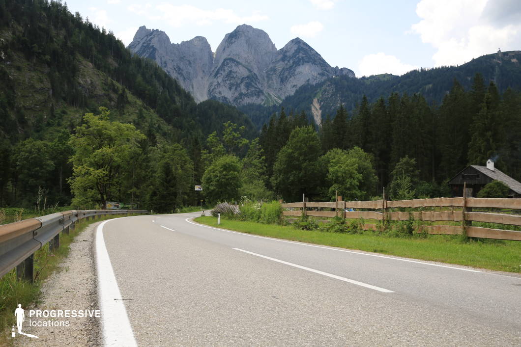 This location features a gently curving asphalt road flanked by metal guardrails on one side and a rustic wooden fence on the other, seamlessly integrating into a densely forested alpine environment. Towering pine trees line the area, while the rugged, grey stone peaks of a mountainous backdrop rise dramatically in the distance, creating a powerful sense of depth and verticality. The color palette is dominated by natural greens and greys, enhancing the landscape's serene yet commanding atmosphere. The textures range from the smooth road surface to the coarse rocky mountain faces and weathered wooden elements, offering rich visual contrast. The perspective is slightly low and angled along the road’s curve, guiding the viewer’s eye into the scene and emphasizing its cinematic composition. This location is ideal for film scenes requiring a secluded, natural setting and works especially well for genres like drama, adventure, or thriller, supporting themes of isolation, transition, or journey.