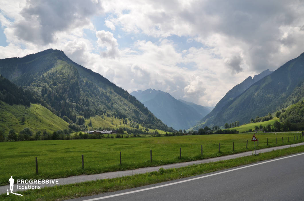 This expansive mountain valley features a broad, open field bordered by gently sloping hills and steep, forested mountain faces that converge in the distance, forming a natural corridor. The dominant green hues of the grass and trees create a harmonious, tranquil landscape, while the interplay of light and shadow on the hillsides adds depth and visual interest. A narrow paved road runs along the foreground, delineated by simple wooden posts and a low roadside barrier, contributing a linear geometric element that contrasts the organic flow of the terrain. The surrounding mountain ridges are covered with dense vegetation, transitioning from lower grassy slopes to high-altitude coniferous forests and rocky peaks. This location’s wide, uninterrupted vista and clear natural layers make it highly suitable for filming scenic transitions, panoramic establishing shots, or introspective sequences in genres such as drama, documentary, or adventure.