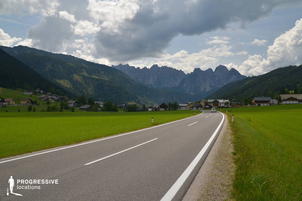 The location features a gently curving asphalt road cutting through a wide, open valley surrounded by rolling hills and distant, sharply contoured mountain peaks. The valley floor is covered by a uniform carpet of vivid green grass, enhancing the clarity and openness of the composition. The buildings in the midground form a compact rural village, their traditional gable roofs and low-rise profiles harmonizing with the natural landscape. Forested slopes frame both sides of the valley, leading the eye toward the jagged, dramatic mountain ridge in the background. The camera captures the scene from road level with a central vanishing point perspective, emphasizing depth and motion. This location is ideal for productions seeking a serene yet expansive rural setting, well-suited for genres such as drama, road movies, or family-oriented storytelling, particularly for scenes requiring scenic travel sequences or wide establishing shots.