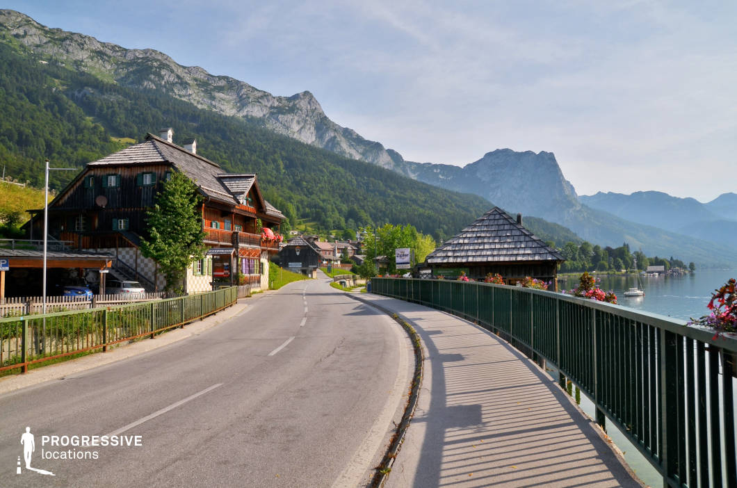 This location presents a gently curving lakeside road flanked by traditional alpine architecture, with dark timber façades, pitched roofs, and flower-adorned balconies adding regional character. The road runs alongside a green metal railing, which separates it from the clear water of the lake on the right, enhancing spatial layering in the composition. On the left, rustic wooden buildings line the slope, leading into a compact village nestled at the foot of steep, forested mountains. The mountainous backdrop offers dramatic natural elevation, while the open sky and long perspective create a visually expansive setting. The dominant color palette includes natural greens, greys, and wood tones, conveying a fresh and harmonious rural ambiance. This location is highly suitable for productions needing an authentic, picturesque village atmosphere with access to water and mountainous scenery, making it ideal for genres such as romance, period drama, or travel-related features, especially for establishing shots or transitional scenes.