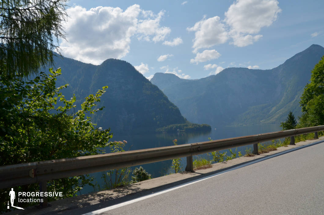 This location features a winding mountain road bordered by a metallic guardrail that runs alongside a deep-blue lake set against a backdrop of steep, forest-covered peaks. The visual composition balances man-made infrastructure and unspoiled natural elements, with the road in the foreground creating a linear contrast to the irregular shapes of the mountainous terrain. The expansive lake surface, framed by the towering slopes, adds depth and calmness to the scene. The mix of rugged rock formations and dense greenery on the mountains contributes to a dramatic, yet serene landscape. Ideal for wide establishing shots, road trip sequences, or introspective moments in natural surroundings, this location lends itself well to drama, adventure, and nature-focused genres.