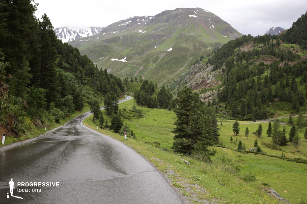 This location features a gently curving asphalt road set within a mountainous landscape, framed by densely forested slopes and scattered rock formations. The road winds into the distance, offering depth and perspective, while the expansive valley on the right side provides a broad visual field. The terrain is characterized by its natural undulations, a mix of grassy surfaces and steep hillsides. Tall coniferous trees dominate the scene, arranged in varied densities, contributing to a rich, evergreen color palette with subtle earth tones from exposed rocky areas. The juxtaposition of the paved infrastructure and the raw natural environment enhances the visual contrast, emphasizing both isolation and connectivity. Ideal for road sequences, travel montages, or serene interludes, this location supports a range of genres including drama, adventure, or nature-focused productions. Its open composition and natural layering also make it suitable for aerial or long-lens cinematography.