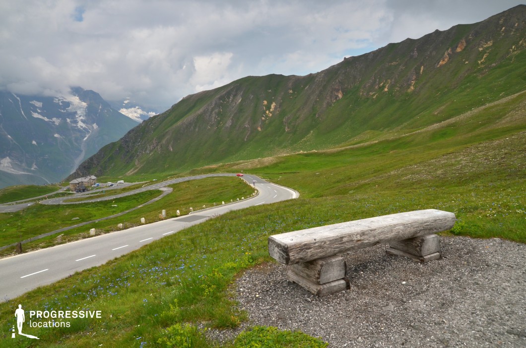 This panoramic mountain location is defined by a gently winding serpentine road cutting through vast green pastures and sloping ridgelines. The foreground features a rough-hewn wooden bench positioned on a gravel clearing, offering a visual anchor and natural resting point that frames the expansive valley view. The grassy hills are dotted with small wildflowers and transition into steeper, rocky elevations that stretch toward a distant, snow-streaked mountain range. The dominant color palette includes vivid greens and muted greys, creating a tranquil yet open ambiance. Shot from a slightly raised perspective, the image captures both the scale and rhythm of the curving road below, making it visually rich and spatially layered. This location is highly versatile for filming, particularly for genres like travel documentaries, introspective dramas, or commercials, with excellent suitability for establishing shots, movement sequences, or reflective scenes in natural, unspoiled surroundings.