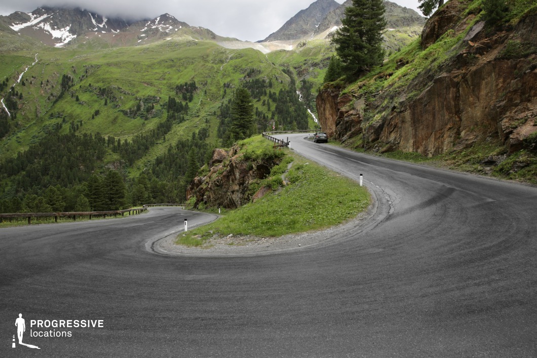 This striking mountain location features a dramatic hairpin turn carved into the slope, bordered on one side by exposed rock faces and on the other by steep drop-offs into a densely wooded valley. The asphalt road surface contrasts with the vivid greenery of the hillside and the reddish-brown rock textures, creating a dynamic visual interplay between natural and man-made elements. The curve itself forms a strong geometric composition, ideal for capturing motion or tension. The surrounding terrain is rugged, with elevated ridges and forested slopes contributing to a layered mountainous backdrop. The elevated camera position emphasizes the depth of the valley and the complexity of the road’s path. This location is particularly suited for action sequences, driving scenes, or dramatic landscape shots in thrillers, adventure films, or commercials requiring a sense of isolation and movement.