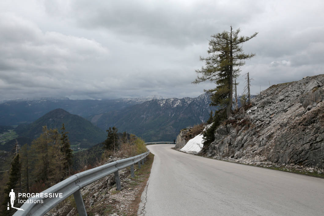 This winding mountain road ascends along a steep, rocky slope, flanked by a rugged guardrail and sparse tree growth emerging from the uneven terrain. The surface is light-toned asphalt, gently curving into the distance, framed on one side by a dramatic drop-off and on the other by coarse rock formations. In the background, expansive views of layered mountain ranges with snow-capped ridges emphasize the road's elevation and remote setting. The sparse vegetation and exposed stone textures evoke a raw, untamed atmosphere. This location is ideal for high-altitude driving sequences, introspective travel scenes, or establishing shots requiring expansive natural vistas, making it suitable for drama, adventure, and documentary-style productions.