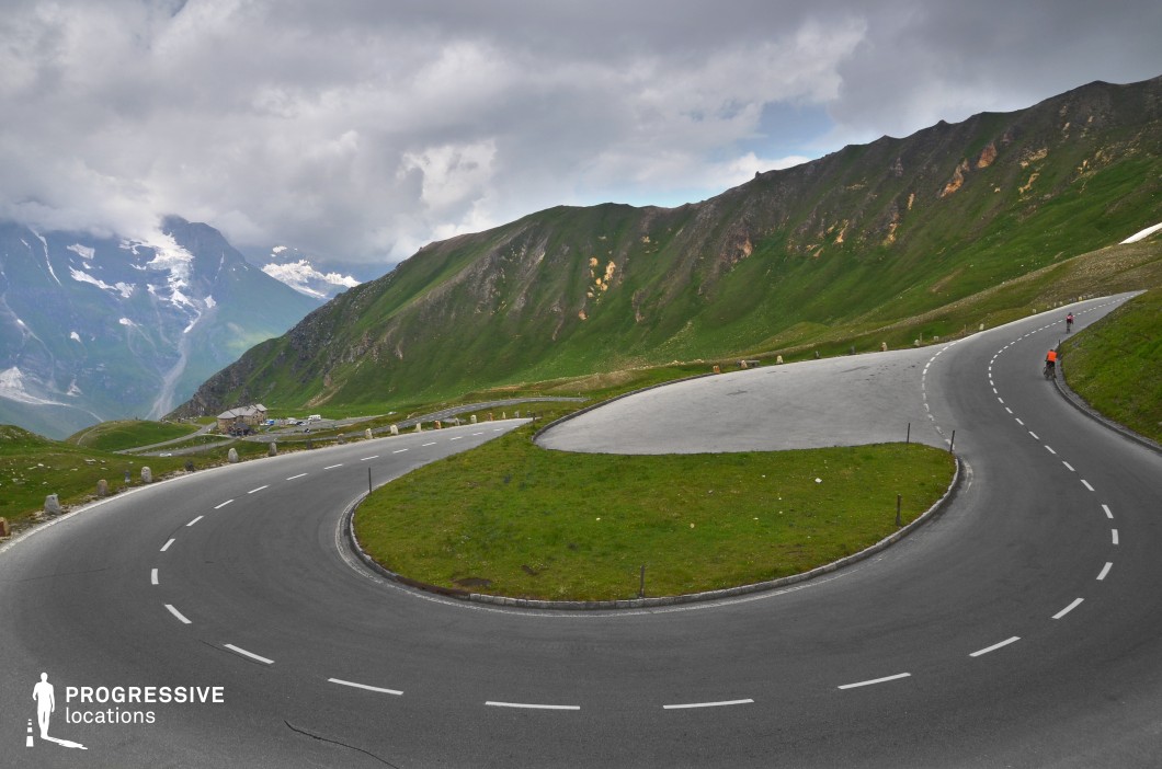This striking location features a dramatic serpentine road cutting sharply through a mountainous alpine landscape. The broad, sweeping hairpin turn dominates the composition, bordered by low stone posts and framed by expansive green slopes and jagged rocky ridges. The paved surface is wide and clean, offering a stark contrast to the surrounding natural textures of grass and exposed stone. The elevation creates strong depth, with distant mountain faces adding scale and grandeur. The visual geometry of the road’s curve enhances cinematic dynamism, making it a compelling site for filming action sequences, dynamic travel scenes, or tension-filled moments in thrillers and adventure genres.
