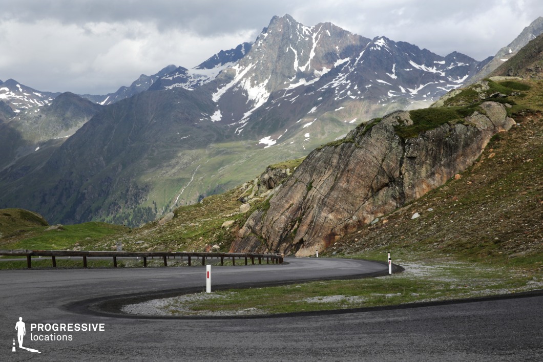 This location presents a striking mountain pass with a sweeping asphalt road curving tightly around a large exposed rock formation. The foreground is dominated by the road’s dark, smooth surface and a low wooden guardrail, while the midground is defined by rugged stone textures and minimal vegetation clinging to the slope. Towering snow-capped peaks rise in the background, forming a dramatic natural skyline with sharp, angular ridgelines. The contrast between the geometric road curvature and the organic forms of the rock and mountain range adds visual dynamism. The scale of the open alpine environment, combined with the road’s distinctive curve, makes this an excellent setting for high-impact driving sequences, outdoor adventure scenes, or establishing shots in genres like action, drama, and thriller. Its expansive visual depth and raw natural setting support both atmospheric and movement-focused cinematography.