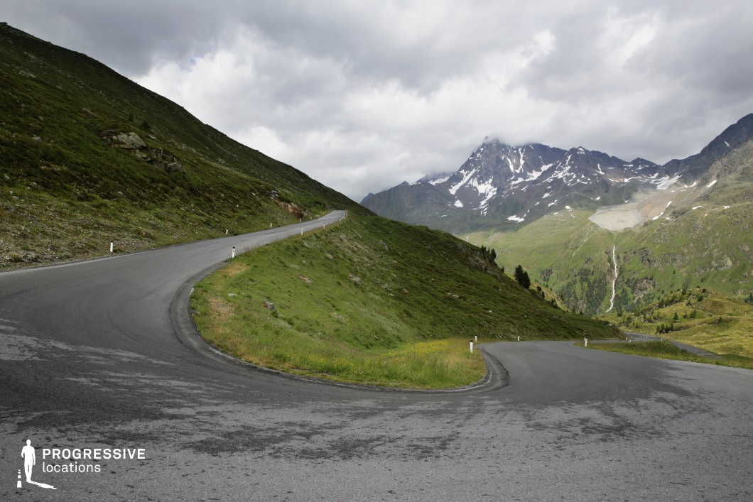 This striking location showcases a steep, serpentine mountain road making a dramatic hairpin turn amid rolling alpine slopes. The dark grey asphalt contrasts sharply with the vivid green of the grassy hillside, creating a bold visual flow that draws the eye upward toward a jagged, snow-patched mountain range in the background. The terrain is predominantly natural, characterized by rugged rock outcrops, sparse alpine vegetation, and a distant cascading waterfall that adds vertical dynamism to the scene. The image is composed from a slightly elevated perspective, emphasizing the road's curvature and depth. The minimal human-made elements and wide-open space enhance its sense of remoteness and scale. This location is especially suited for filming high-altitude sequences in genres such as action, thriller, or drama, and is ideal for showcasing themes of challenge, solitude, or movement through isolated terrain.