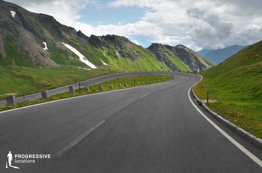 A sweeping curve of freshly paved asphalt carves through an expansive alpine pass, surrounded by vibrant green slopes and jagged mountain ridges. The road is visually emphasized by a series of evenly spaced stone markers and a distinct white edge line, drawing the eye along its graceful arc. The mountainous terrain combines exposed rocky textures and grassy patches, with isolated snow remnants adding contrast to the vivid natural palette. The open perspective and rhythmic road design enhance depth and dynamism within the frame. This location offers a cinematic setting for dynamic driving scenes, introspective travel sequences, or sweeping landscape visuals, ideal for road movies, adventure genres, and commercials emphasizing movement and freedom.