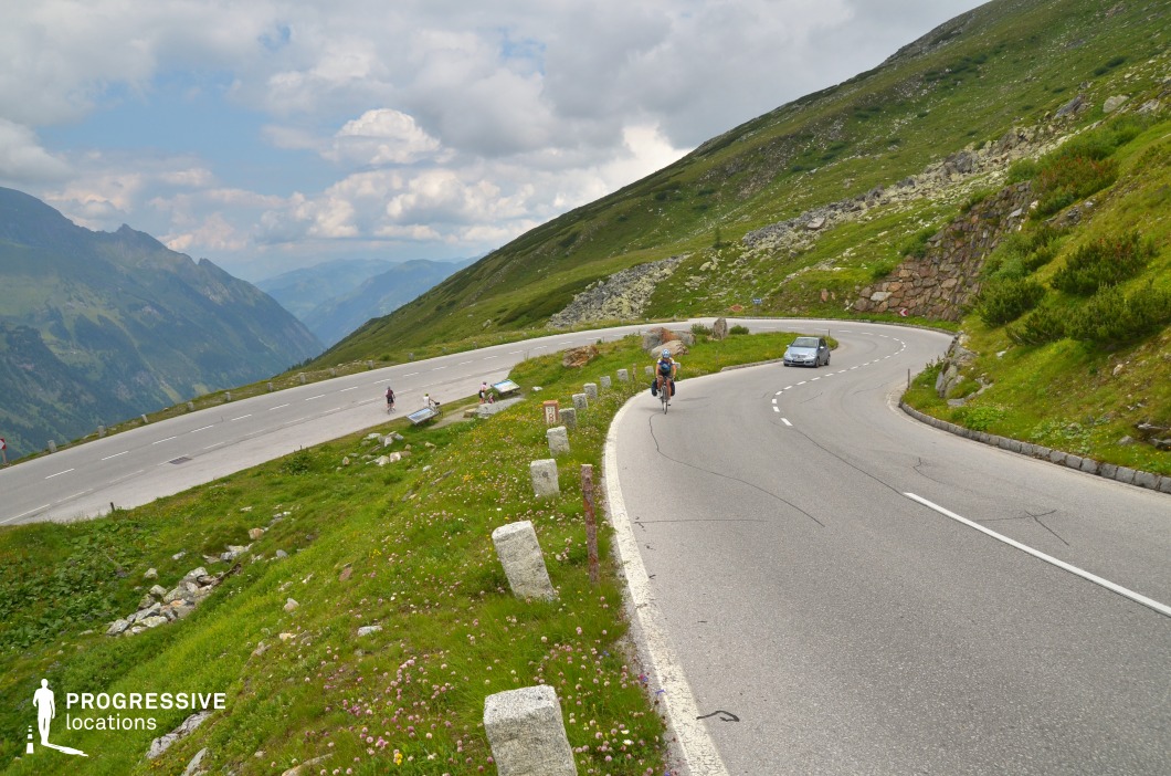 This mountain pass features a wide, well-maintained asphalt road winding uphill across a lush alpine slope. The road curves gently against a backdrop of expansive green hills dotted with rocky outcrops and low-lying vegetation, with steep drops on one side and a rising slope on the other. The roadside is bordered by evenly spaced stone markers, enhancing the location’s sense of structure and rhythm. The horizon opens to a vast mountain range layered in the distance, adding depth and perspective to the scene. The bright, natural color palette—dominated by greens and greys—emphasizes the freshness and clarity of the high-altitude setting. This location is ideal for filming in genres such as adventure, documentary, or sports-focused projects, particularly for scenes involving movement, elevation, and vast natural panoramas. Its clean layout and scenic layering support aerial, tracking, or dynamic wide shots.