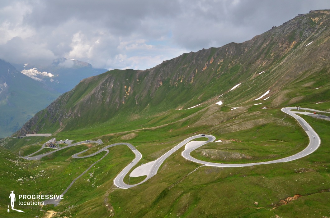This high-angle view reveals a wide, looping serpentine road intricately embedded in a sweeping alpine landscape. The undulating terrain is blanketed in vivid green vegetation, dotted with patches of exposed rock and residual snow, emphasizing the altitude and natural texture of the environment. The road's sinuous geometry creates a striking contrast against the organic contours of the mountains, providing visual rhythm and motion across the frame. The broad curves and openness of the valley allow for expansive camera movements and clear line-of-sight, making the location ideal for aerial shots, chase scenes, or scenic transitions in genres such as action, drama, or documentary.