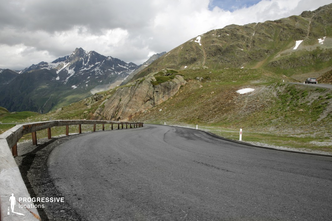This sharply curving mountain road is set within a rugged alpine environment, bordered by a wooden guardrail and flanked by grassy slopes and exposed rock formations. The road’s smooth, dark surface contrasts with the irregular textures of the surrounding terrain, while distant snow-streaked peaks anchor the backdrop with dramatic verticality. The geological variety—ranging from compacted gravel to large boulders and sloping ridgelines—adds depth and complexity to the visual structure. With its open sightlines, elevation, and sense of isolation, this location is ideal for dynamic driving sequences, tension-building transitions, or landscape-focused visuals, making it well-suited for adventure, thriller, or automotive-themed film projects.