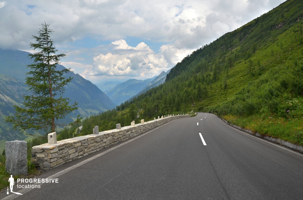 This high-altitude road winds along a steep, forested mountainside, bordered by a low stone guardrail composed of light-colored stacked blocks that add a crafted, textural element to the scene. The road surface is smooth and freshly marked, leading the eye into a vast panoramic view of layered mountain ridges in the background. The left side drops into a deep valley, creating a strong sense of verticality, while the right side rises into dense green foliage. The subtle curvature of the road and the linear perspective accentuate motion and depth, offering excellent visual rhythm. This setting is ideal for dynamic driving scenes, cinematic landscape reveals, or commercials, and fits well within genres like drama, nature documentaries, or adventure films due to its expansive and visually balanced composition.