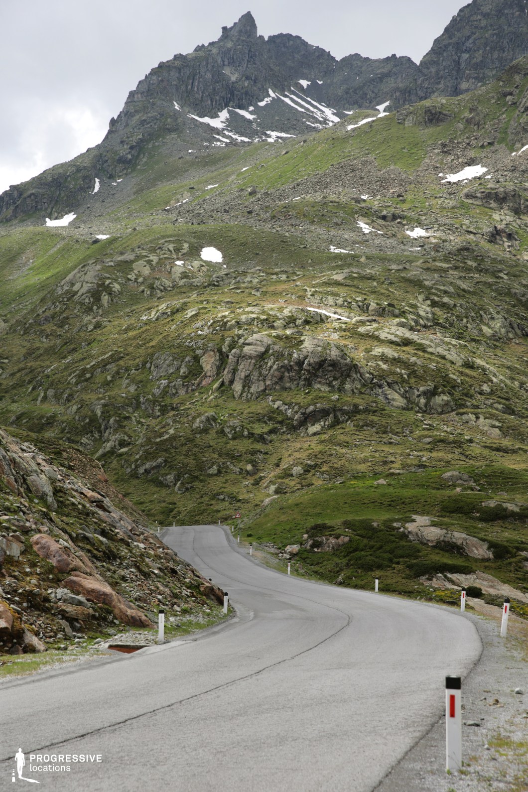 This rugged mountain setting is dominated by jagged peaks and a barren, rocky terrain interspersed with patches of moss and low vegetation. The narrow, winding road in the foreground ascends through a raw alpine landscape, flanked by steep rock formations and scattered boulders. The grey asphalt contrasts starkly with the irregular green and brown tones of the mountainside, while the sparse snow remnants on the higher ridges hint at the elevation and isolation of the environment. The vertical framing emphasizes the towering cliffs above, creating a sense of dramatic scale and harsh natural grandeur. This visually striking, desolate mountain pass is ideal for filming high-altitude travel scenes, remote wilderness narratives, or tension-filled sequences in thrillers and survival-themed productions.