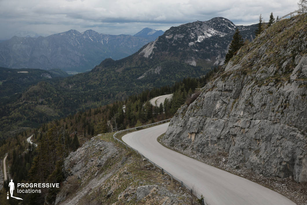This high-altitude mountain road winds along a steep cliff face, offering sweeping views across a vast, forested valley and distant mountain ranges. The light-colored asphalt contrasts with the rugged, vertical rock wall it curves around, creating a striking visual interplay between human-made structure and natural geology. The switchback road, bordered by safety rails, introduces strong horizontal and serpentine lines that guide the eye through the layered composition of trees, slopes, and distant peaks. The rocky surface is harsh and textured, enhancing the dramatic and raw aesthetic of the location. Captured from an elevated perspective, the scene emphasizes elevation and vastness. This location is highly suitable for filming dramatic sequences, scenic drives, or adventurous transitions in genres like action, travel, or nature documentaries, where the expansive views and dynamic terrain provide both visual tension and cinematic depth.