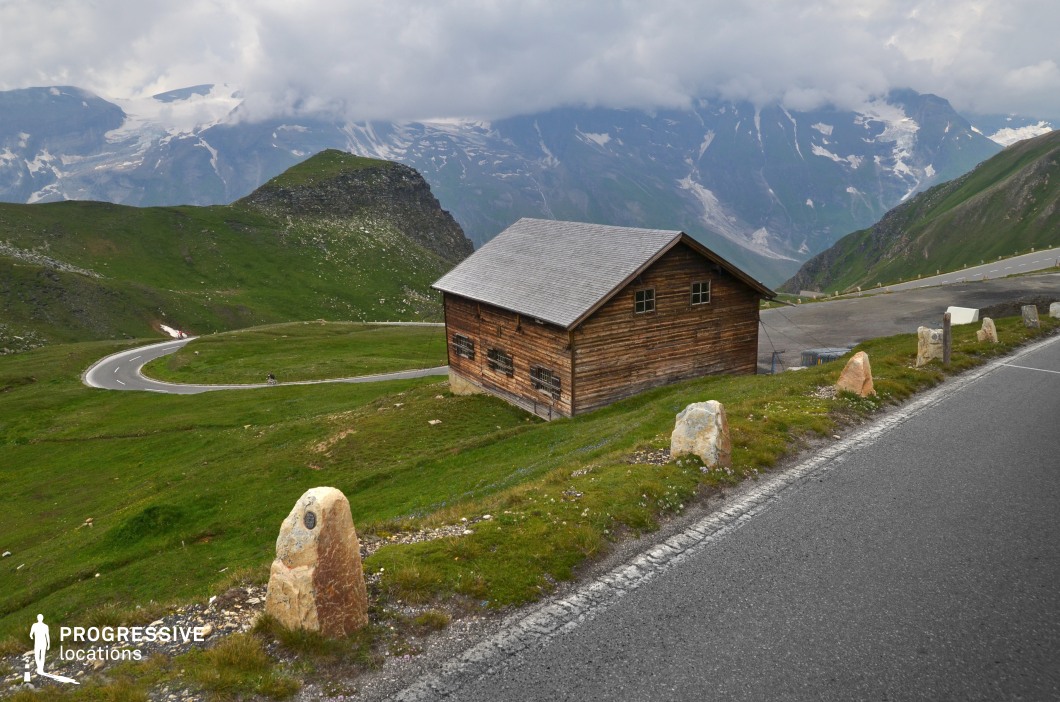 This elevated mountain location presents a panoramic view with gently curving roads, rolling green hills, and a solitary wooden cabin positioned prominently in the midground. The cabin, constructed from weathered timber with a simple gable roof, introduces a rustic architectural element that contrasts with the expansive natural surroundings. The terrain is composed of open grassy surfaces interspersed with rocky outcrops, and the winding road enhances the scene's sense of movement and spatial depth. The distant backdrop of snow-patched peaks and steep cliffs adds dramatic scale and layered composition. The site’s open layout, architectural focus, and natural vistas make it particularly well-suited for landscape-driven productions, reflective sequences, or genre scenes in drama, historical narratives, or travel-oriented content.