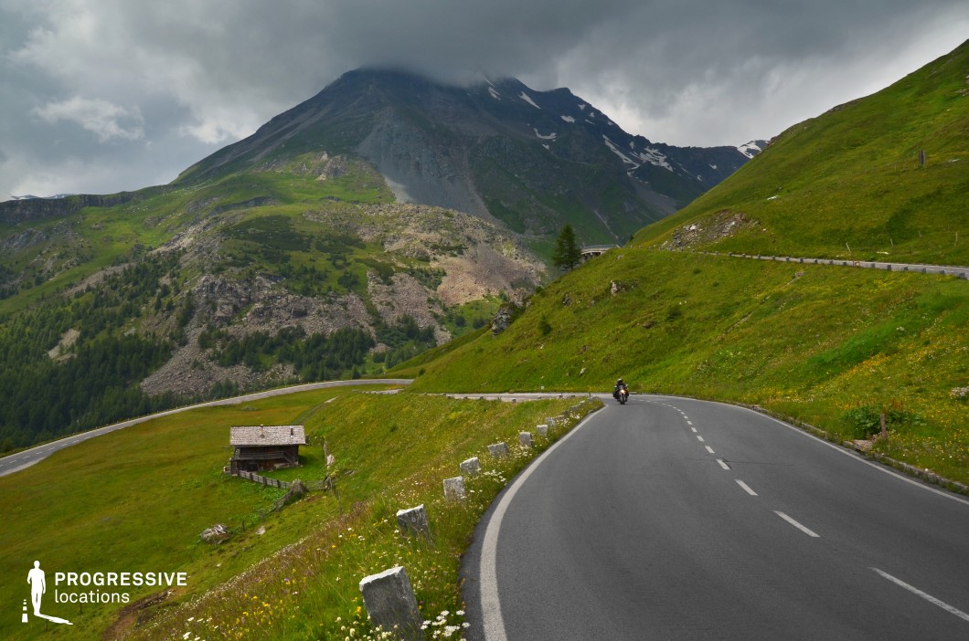 This scene showcases a winding mountain road that cuts through rolling alpine meadows, framed by steep green hills and a towering rocky peak in the background. The paved road features clean white lane markings and a row of low stone barriers, emphasizing its gentle incline and safe structure. A rustic wooden cabin sits lower on the slope, adding architectural contrast to the expansive, untouched landscape. The terrain is layered with rocky outcrops, scattered vegetation, and a mix of grassy textures, enhancing the visual complexity of the setting. The backdrop includes a dramatic mountain partly obscured by low-hanging cloud cover, contributing to the sense of elevation and scale. This location’s open, curving road and layered geography make it ideal for cinematic tracking shots, travel sequences, or motorcycle and driving scenes in adventure, drama, and road movie genres.