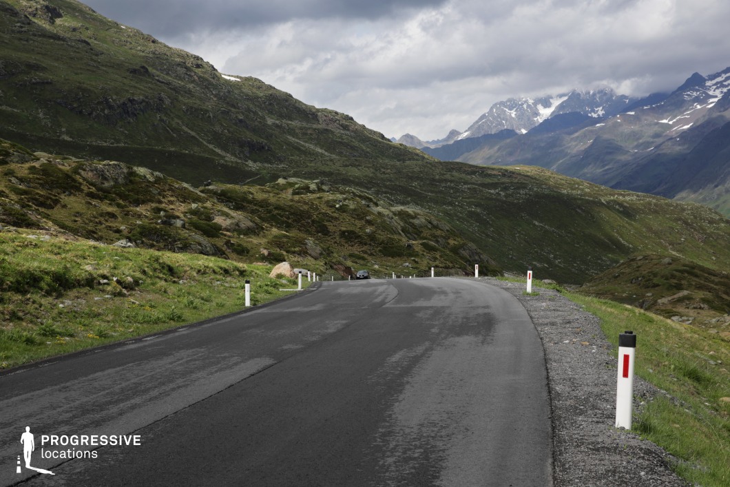 This remote mountain road curves gently through a rugged alpine environment characterized by sloping green hills, rocky outcrops, and distant snow-capped peaks. The asphalt surface is dark and subtly reflective, bordered by gravel shoulders and simple reflective road markers, emphasizing functional minimalism. The terrain rises and falls dramatically, with the surrounding landscape offering a layered composition of rolling hills in the foreground and jagged ridgelines in the background. The openness of the perspective and the expansive natural backdrop lend the scene a strong sense of scale and isolation. This location is well-suited for film productions needing solitary, high-altitude settings for scenes in genres such as thrillers, road movies, or introspective dramas.