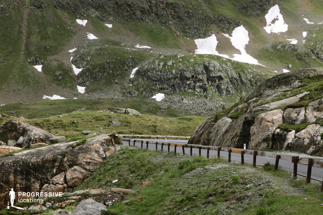 This image captures a narrow, curving asphalt road that cuts through a rugged alpine landscape, flanked by large, weathered boulders and grassy slopes. The scene is dominated by a dramatic interplay of textures: coarse stone surfaces, sparse alpine vegetation, and patches of lingering snow across the steep, uneven mountainsides. The road itself is visually accentuated by its dark surface and metal barrier, winding through the natural formations in a way that emphasizes the isolation and scale of the environment. With its untamed topography and raw, elemental aesthetic, this location is highly suitable for scenes requiring a sense of remoteness or tension, fitting well for thriller, action, or exploration-themed productions.