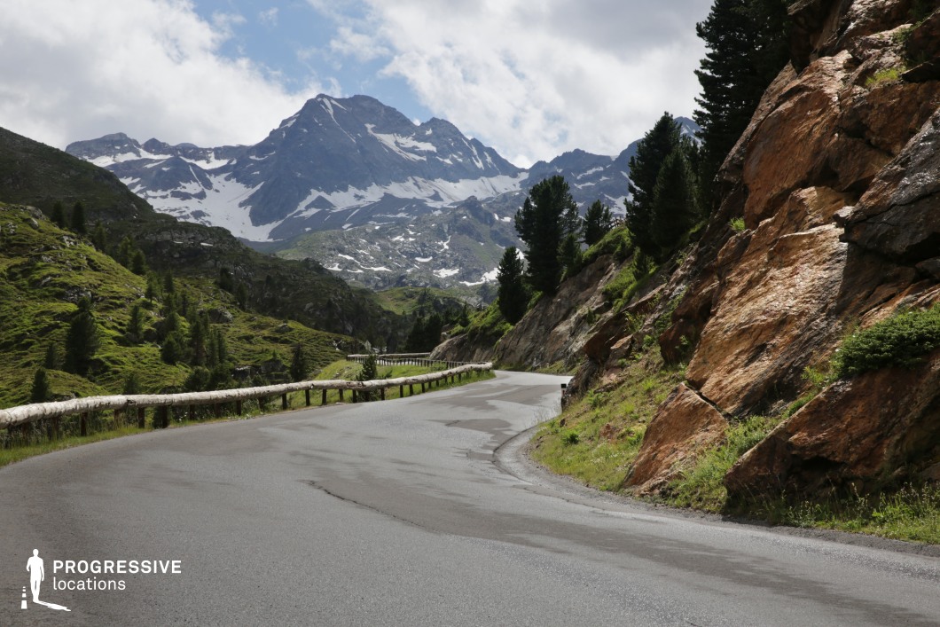 This mountain road curves gracefully through a high-altitude alpine environment, bordered on one side by rugged, reddish rock formations and on the other by a wooden guardrail overlooking a steep, grassy valley. In the background, a dramatic snow-patched peak dominates the skyline, contributing to a striking sense of scale and elevation. The exposed rock surfaces introduce bold, textured elements that contrast with the soft greenery and darker pine trees scattered along the slopes. The clean, well-maintained road surface and serpentine alignment provide a dynamic leading line through the scene, enhancing its cinematic depth. This location’s distinctive geological textures, high-contrast terrain, and unobstructed views make it ideal for filming travel sequences, adventure scenes, or commercial productions set in remote or majestic landscapes.