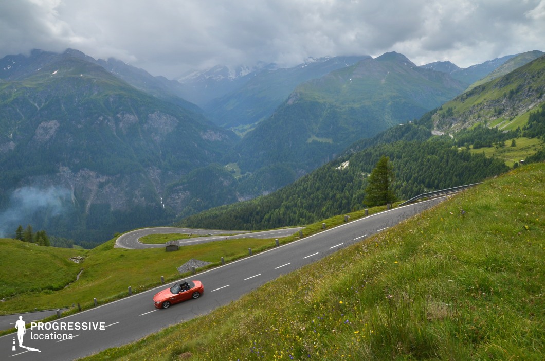 This elevated mountain location features a steep, winding road that zigzags through densely forested slopes and open alpine meadows, with a sweeping view over vast, pine-covered valleys and distant layered ridges. The midground showcases a dramatic hairpin curve, visually anchoring the composition and emphasizing the topography’s steep incline. Dominated by green hues across varying textures—rocky outcrops, grassy embankments, and dense coniferous vegetation—the landscape offers a cohesive and immersive natural setting. The wide view and long sightlines enhance spatial depth, while the smooth asphalt roadway provides a modern contrast to the surrounding rugged environment. This visually dynamic location is ideal for filming action sequences, car commercials, or travel-related productions where scale, elevation, and motion are essential elements.