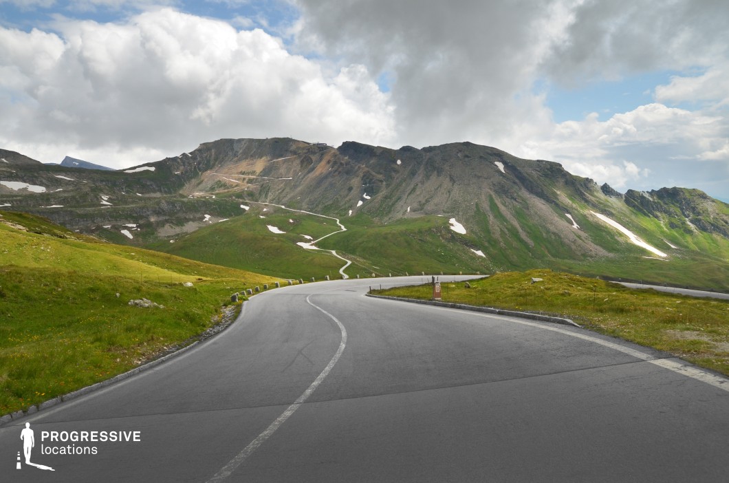 This striking mountain location is defined by a broad, dark asphalt road curving smoothly through an elevated, open alpine landscape. The gentle green slopes are dotted with rocky outcrops and patches of snow, providing a natural texture contrast that adds depth to the scene. The distant mountain ridge exhibits exposed rock surfaces and sharp contours, enhancing the dramatic terrain geometry. The road features minimal barriers and follows a meandering path visible far into the distance, emphasizing scale and motion potential. The wide perspective and clean composition make this location highly adaptable for cinematic road scenes, vehicle shots, or panoramic transitions, particularly in adventure, drama, or travel-related productions.