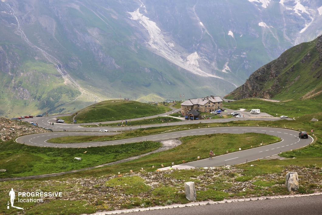 This mountain pass location features a striking serpentine road layout that carves its way through a lush green landscape, offering dynamic lines and curves ideal for visual storytelling. A prominent stone lodge with a slate roof sits centrally in the composition, constructed in a traditional alpine style that reinforces the high-altitude setting. The surrounding terrain is composed of grassy ridges and rugged slopes, with distant cliffs and rocky surfaces providing a dramatic backdrop. The elevated vantage point allows for a wide, layered view of the environment, creating strong compositional depth. The scene’s combination of architectural presence, complex road geometry, and expansive vistas makes it especially suitable for car commercials, cycling sequences, or narrative films in genres such as drama, adventure, or travel documentaries.